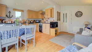 A compact kitchen with wooden cabinets, a dining table with chairs, a TV, and a sofa in a living area. - 2 Bed in Padstow (oc-p00551) (Penrose)