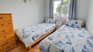 A bedroom with two single beds, a wooden dresser, a nightstand, and a window with curtains. - 2 Bed in Padstow (oc-p00551) (Penrose)