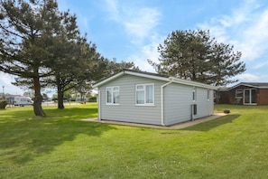 A small, single-story mobile home with a flat roof and a front porch. It is surrounded by a grassy lawn and trees. - 2 Bed in Padstow (oc-p00551) (Penrose)