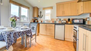 A kitchen with wooden cabinets, a dishwasher, a stove, and a dining area with a table set for two. - 2 Bed in Padstow (oc-p00551) (Penrose)