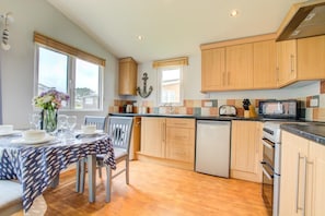 A kitchen with wooden cabinets, a dishwasher, a stove, and a dining area with a table set for two. - 2 Bed in Padstow (oc-p00551) (Penrose)