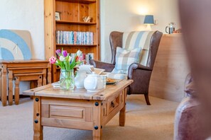 A cozy living room with a wooden coffee table, a bookshelf, a comfortable armchair, and a lamp. - 1 Bed in Padstow (oc-p00554) (Saint Merryn)