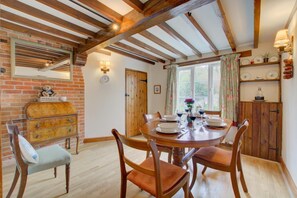 A dining room with a wooden table set for four, surrounded by wooden chairs. A brick wall and a wooden cabinet with dishes are visible. - 2 Bed in Happisburgh (oc-1634) (Happisburgh)