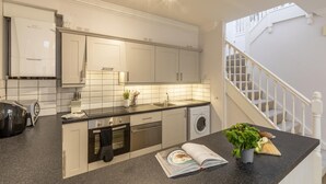 Una cocina moderna con armarios blancos, horno empotrado, lavadora y encimera con un libro y una planta.