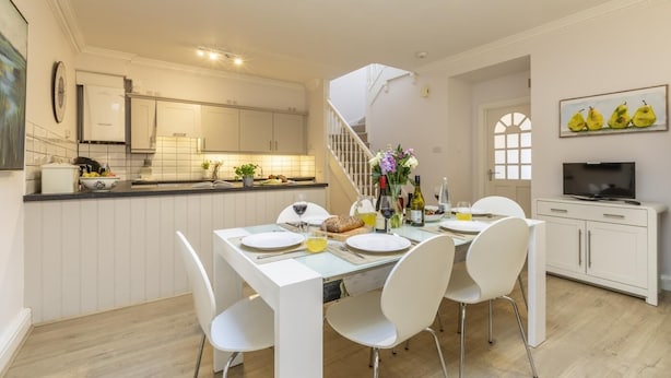 Una cocina moderna con armarios blancos, un comedor con sillas blancas y una escalera que sube a otro nivel.