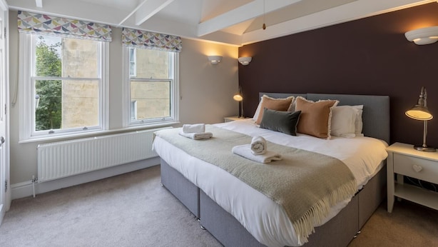 A bedroom with a large bed, two bedside lamps, a radiator, and a window with patterned curtains. - 2 Bed in Bath (oc-bhr24) (Bath)