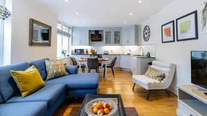 A modern living room with a blue sofa, a white armchair, and a coffee table with fruit. There is a kitchen in the background with white cabinets and a built-in oven. - 2 Bed in Bath (oc-bhr24) (Bath)