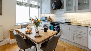 A modern kitchen with white cabinetry, a wooden dining table, and a window with patterned curtains. - 2 Bed in Bath (oc-bhr24) (Bath)