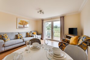 A living room with a grey sofa, yellow and grey patterned cushions, a dining table set with plates and glasses, and a television on a stand. - 3 Bed in Wangford (oc-75sare) (Wangford)