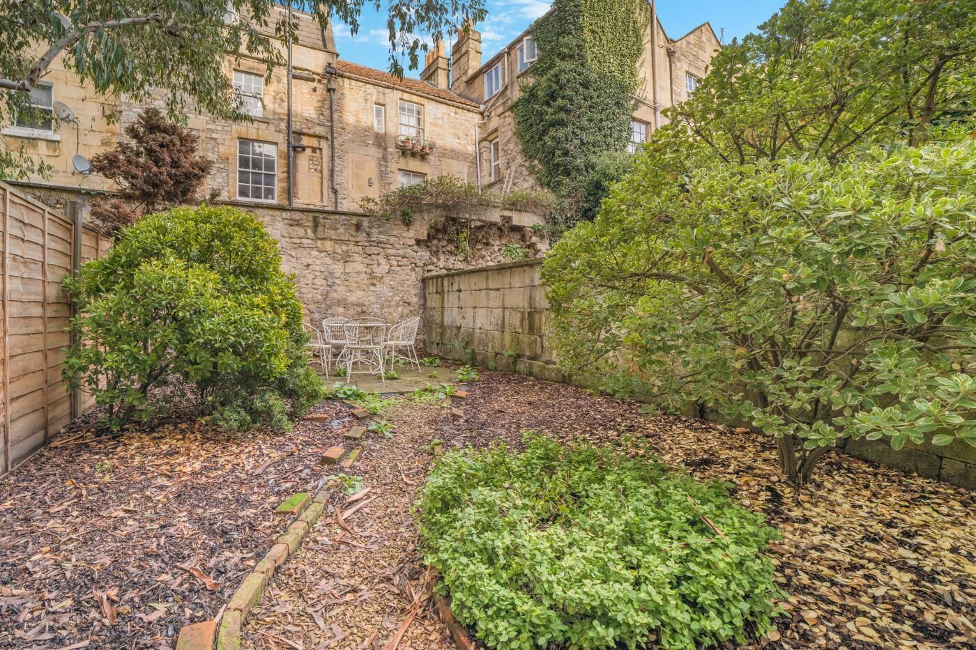 A courtyard with stone walls, a white metal table and chairs, and various shrubs and trees.