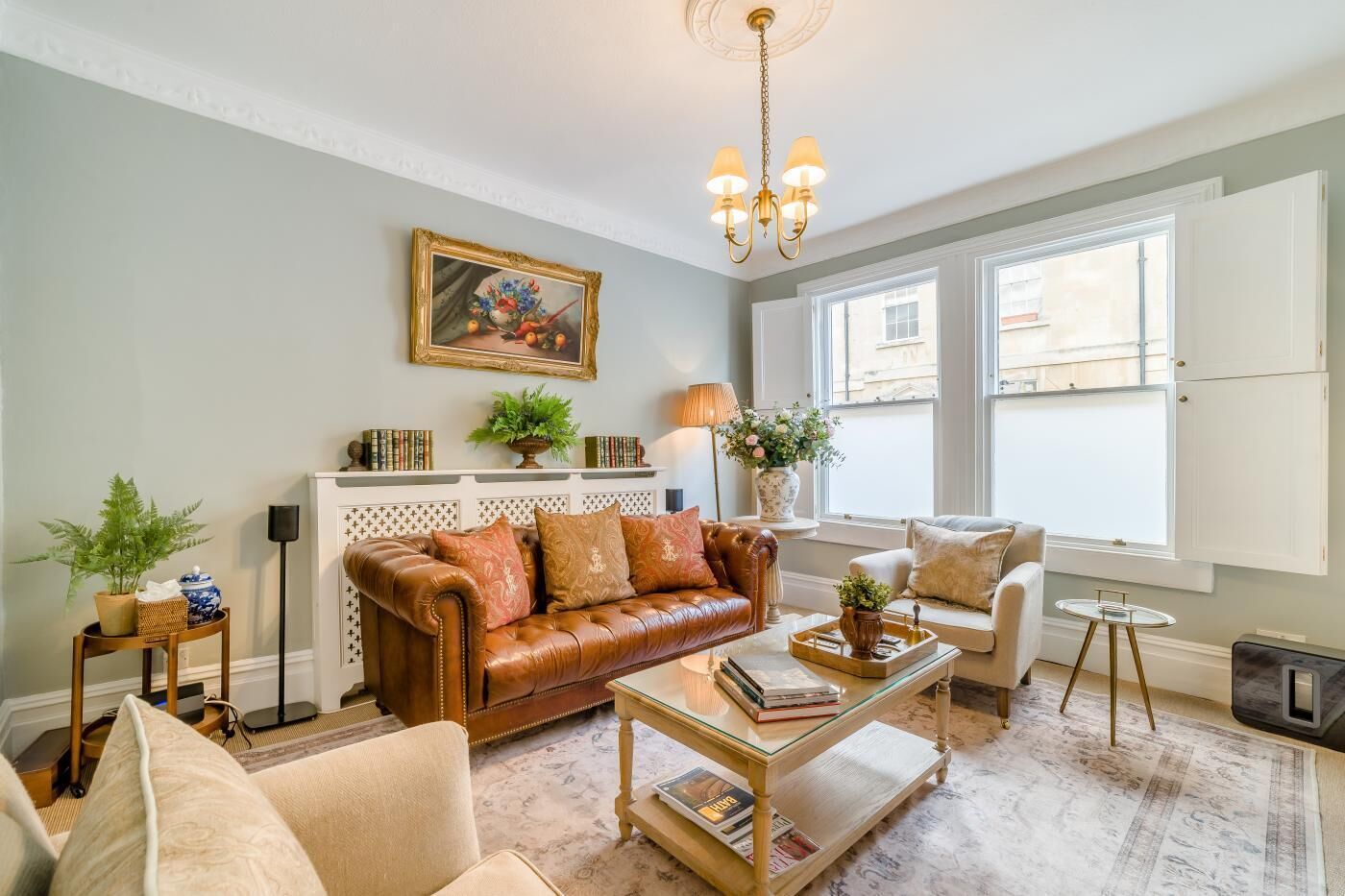 Una sala de estar con un sofá de cuero marrón, una mesa de centro de vidrio, un sillón beige y una pequeña mesa redonda. Hay una estantería con libros y objetos decorativos, un cuadro en la pared y un candelabro.