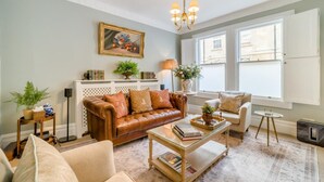 A living room with a brown leather sofa, a glass coffee table, a beige armchair, and a small round table. There is a bookshelf with books and decorative items, a painting on the wall, and a chandelier. - 2 Bed in Bath (oc-bhr25) (Bath)