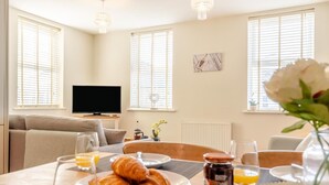 A dining area with a table set for breakfast, including croissants and orange juice, in a room with a television, windows, and a radiator. - 2 Bed in Seahouses (oc-nb1944) (Seahouses)