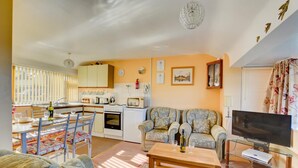 Ein gemütliches Wohnzimmer mit einer Couch, einem Couchtisch und einem Fernseher. Es gibt einen Essbereich mit Tisch und Stühlen sowie eine Küche mit Herd und Mikrowelle.