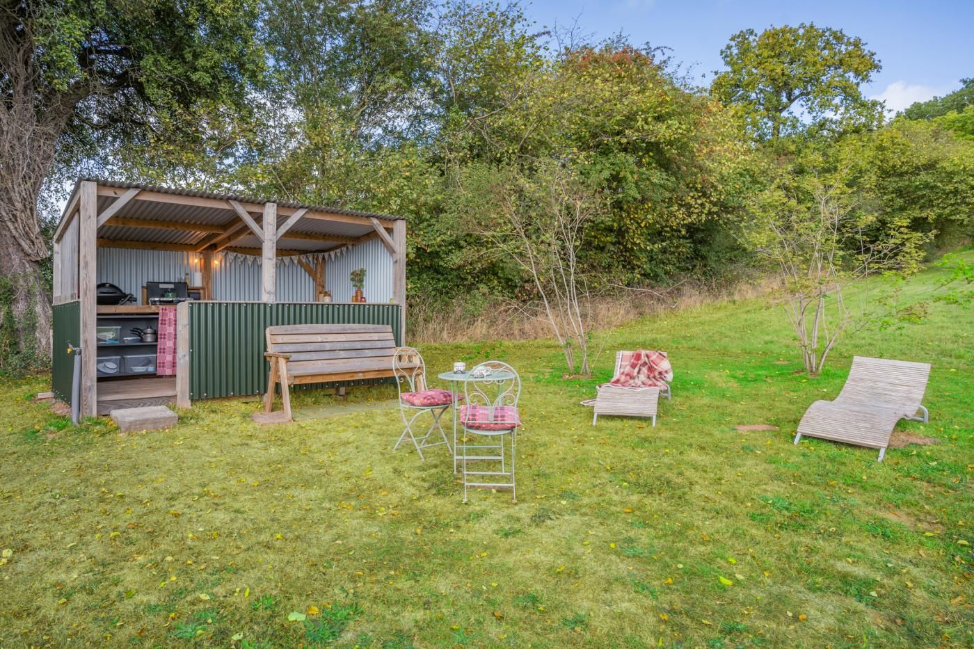 A rustic outdoor kitchen with a wooden bench, two metal chairs, and a table with a plaid tablecloth.