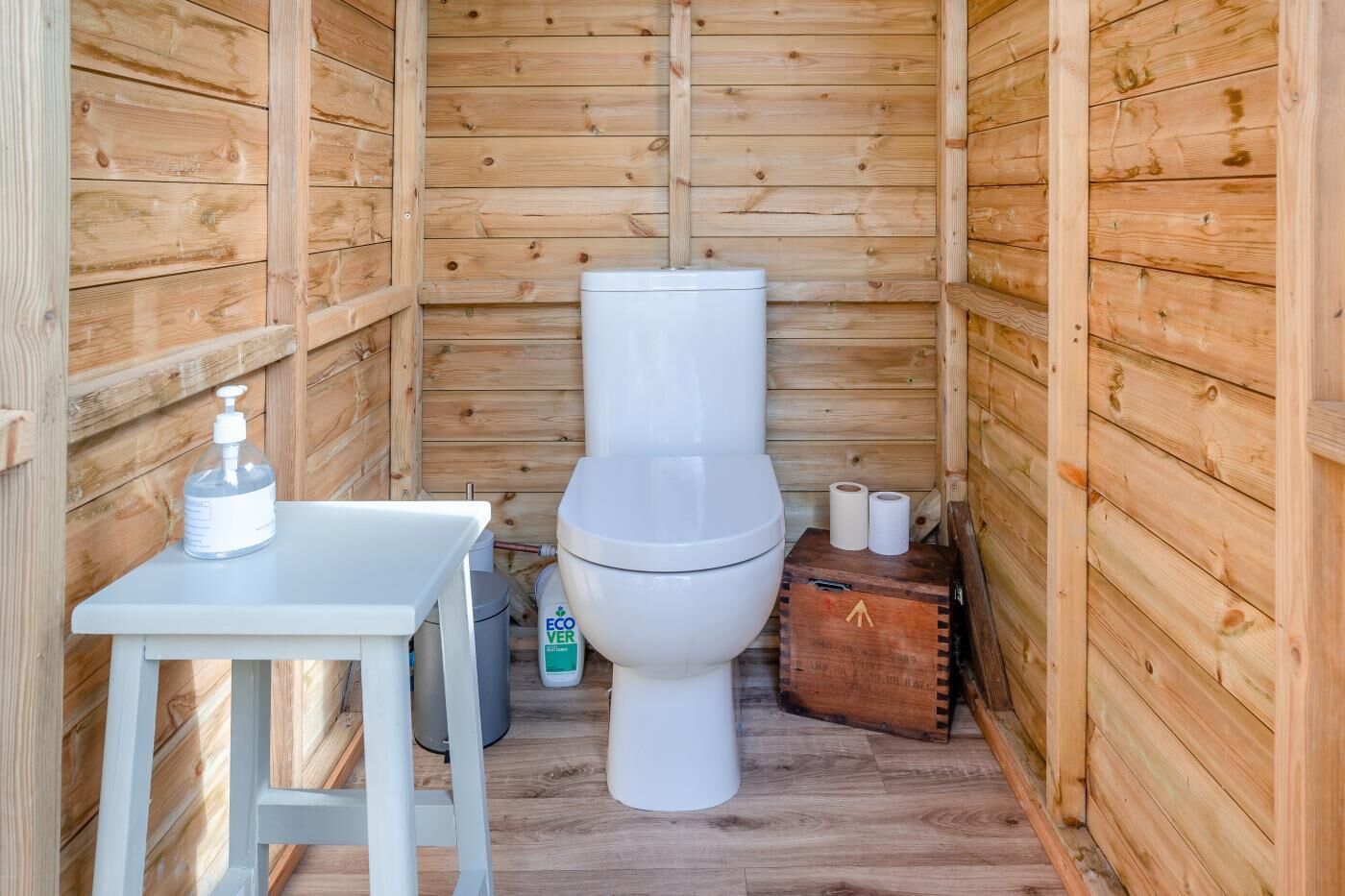 A small wooden cabin with a white toilet, a small table with a soap dispenser, and a trash bin.