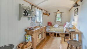 Um quarto aconchegante com uma cama de madeira, uma pequena escrivaninha, uma cadeira, um cesto de lenha e uma prateleira com utensílios de cozinha.