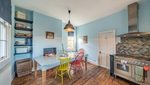 A kitchen with a dining table, chairs, a hanging light fixture, a stove, and shelves with kitchen items. - 3 Bed in Bath (oc-bhr26) (Bath)