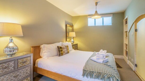 A neatly made bed with white linens, a checkered throw blanket, and a pair of folded towels. There is a bedside lamp, a nightstand with a decorative lamp, and a wall-mounted mirror. - 3 Bed in Bath (oc-bhr26) (Bath)