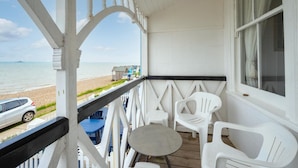 Eine überdachte Veranda mit weißen Geländern, einem kleinen Tisch und zwei weißen Plastikstühlen. Im Hintergrund ist ein Auto am Strand geparkt.