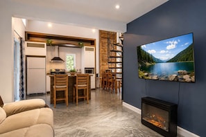 A modern living room with a fireplace, a dining area with wooden chairs, a kitchen with a built-in stove, and a large TV on the wall. - Chalet Chic et Spa | St-Come (Saint-Côme)