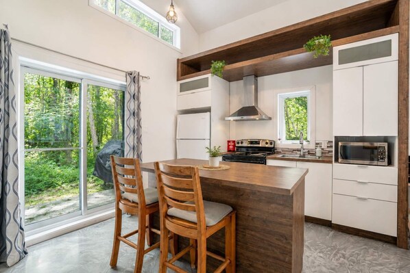 A modern kitchen with a central island, wooden chairs, a stove, a microwave, and a large window with a view of greenery. - Chalet Chic et Spa | St-Come (Saint-Côme)