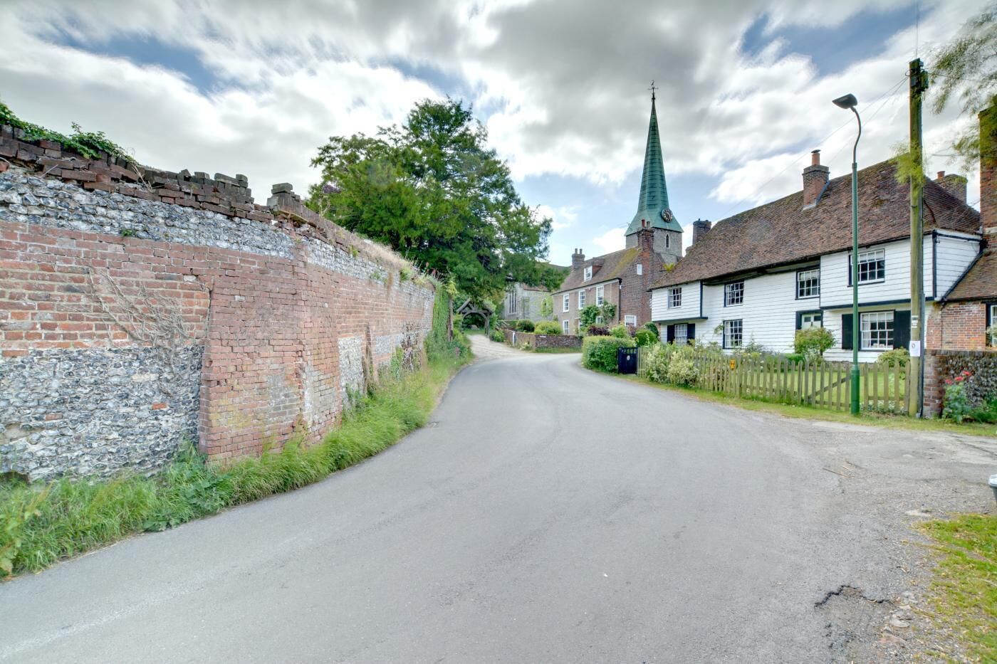 Une rue étroite bordée d’un mur de pierre et de bâtiments traditionnels, dont une église coiffée d’un clocher vert.
