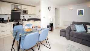Una cocina moderna con armarios blancos, encimera negra y electrodomésticos de acero inoxidable. Hay una zona de comedor con mesa de vidrio y sillas azules. En la sala de estar hay un sofá gris y una lámpara blanca.
