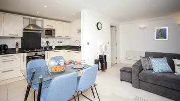A modern kitchen with white cabinets, a black countertop, and stainless steel appliances. There is a dining area with a glass table and blue chairs. A gray sofa and a white lamp are in the living area.