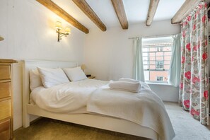 A bedroom with a bed, wooden headboard, bedside lamp, window with curtains, and a view of buildings outside.