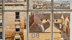 Une vue à travers une fenêtre à carreaux, qui révèle une scène de rue avec différents bâtiments, notamment des maisons d’habitation et un bâtiment plus grand doté d’un toit en tuiles.