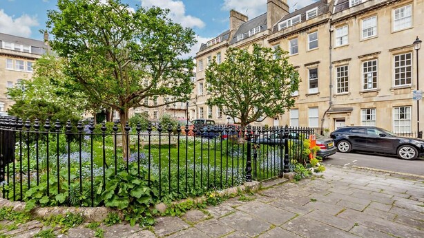 Une rue résidentielle bordée de maisons mitoyennes, avec une clôture en métal noir et un jardin où poussent différentes espèces de plantes.