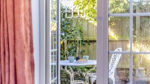 A view through a glass door of a patio area with a table set for tea, surrounded by a wooden fence and greenery. - 1 Bed in Stiffkey (oc-1642) (Stiffkey)