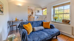A cozy living room with a blue sofa, two yellow cushions, a dining table with chairs, a kitchen area with white cabinets, and a large window with a view of greenery. - 1 Bed in Stiffkey (oc-1642) (Stiffkey)