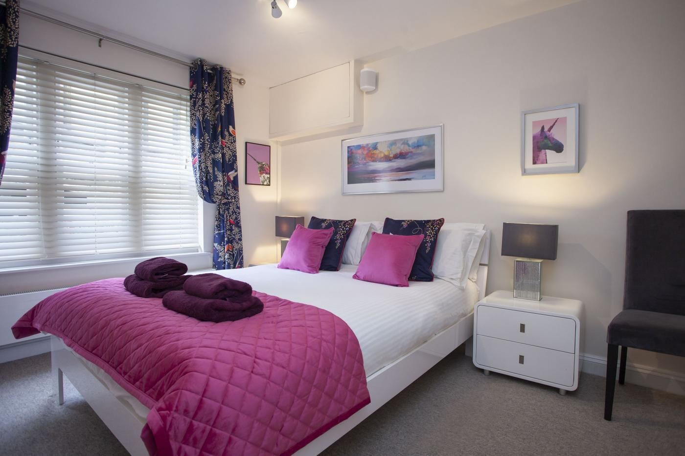 A bedroom with a bed, bedside tables, a nightstand, a chair, and artwork on the walls.