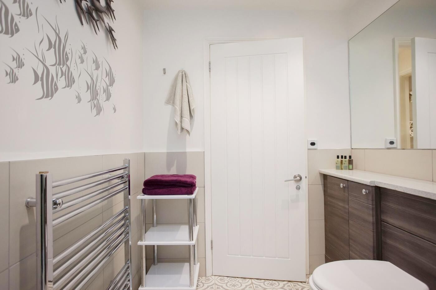 Ein Badezimmer mit einer weißen Tür, einem Handtuchhalter, einem Regal mit einem violetten Handtuch, einer Toilette und einem Spiegel über dem Waschbecken.