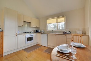 A modern kitchen with white cabinets, a stove, microwave, and dishwasher. There is a dining table with plates, cutlery, and a glass. - 2 Bed in Attleborough (oc-1277) (Old Buckenham)