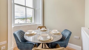 Comedor con una mesa redonda dispuesta para cuatro personas, dos sillas tapizadas en azul y un ventanal con vistas a casas.