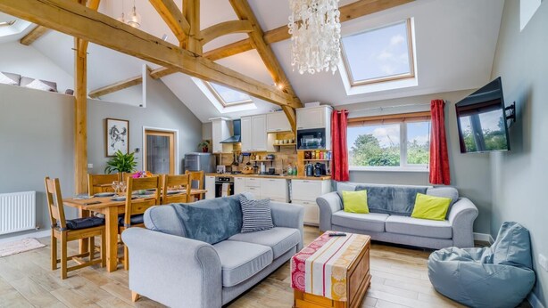 Een ruime living in loftstijl met een hoge plafond, houten balken, een eettafel, banken en een keuken op de achtergrond.