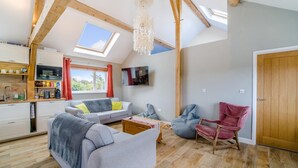 Een ruime woonkamer met een hoge plafond, houten balken, een bank, een bijzettafeltje en een aan de muur bevestigde televisie.