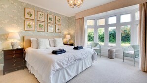 A bedroom with a large bed, two bedside tables with lamps, a chandelier, a window with curtains, and framed botanical prints on the wall. - 2 Bed in Binham (oc-1477) (Binham)