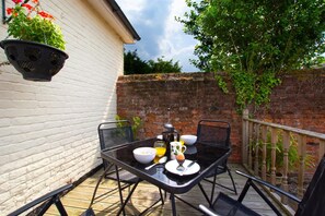 A small outdoor patio with a table set for two, a hanging planter with flowers, and a brick wall in the background.