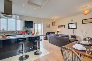 A modern kitchen with a central island, a dining area with a table and chairs, a sofa, and a balcony with a view. - 1 Bed in Blofield Heath (oc-1468) (Blofield)
