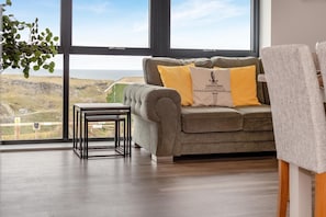 A room with a large window, a grey sofa with yellow cushions, a small table, and a plant. - 3 Bed in Tintagel (oc-e32938) (Tintagel)