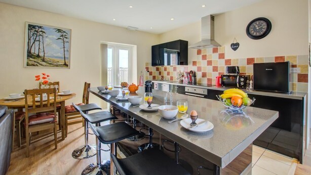 A modern kitchen with a central island, bar stools, a dining table, and a painting on the wall. - 2 Bed in Blofield Heath (oc-1467) (Hemblington)