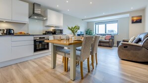 Een moderne keuken met witte kastjes, een eethoek met een houten tafel en stoelen, een bank en een relaxfauteuil.