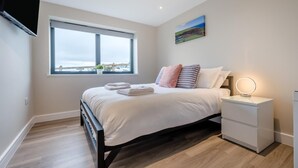Een moderne slaapkamer met een groot raam, een bed met witte lakens, een nachtkastje met een lamp, en een schilderij aan de muur.