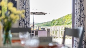 Ein Essbereich mit Tisch für zwei Personen, einem Korbstuhl und Blick auf eine Holzterrasse und Hügel.