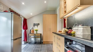 A modern kitchen with a refrigerator, wooden cabinets, a countertop with fruits, and a table with flowers. - 1 Bed in Llandeloy (oc-c32808) (Llandeloy)