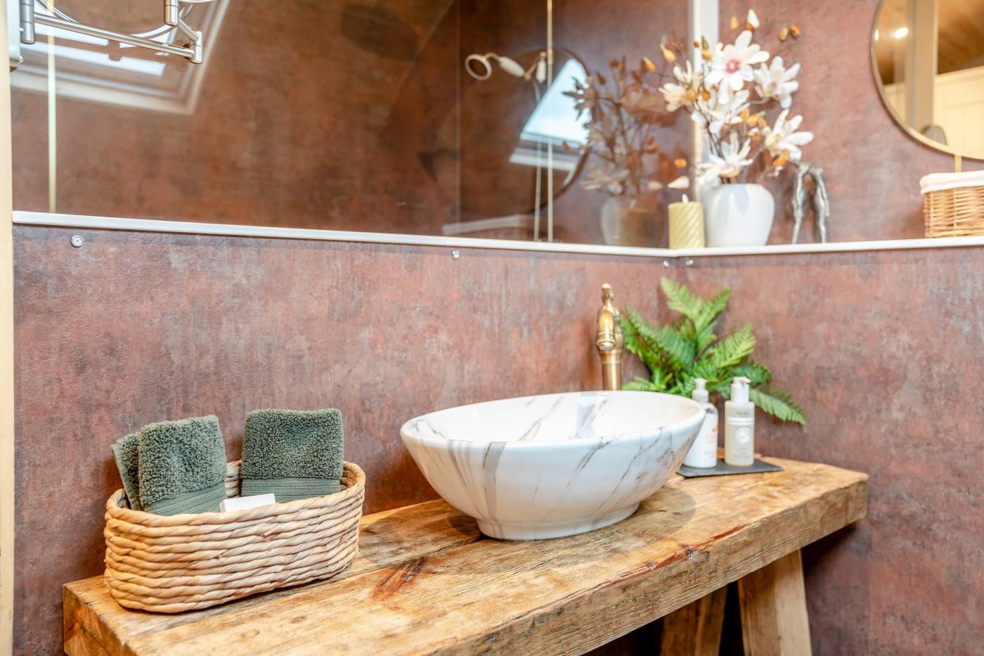A bathroom with a marble sink, a wooden vanity, a woven basket with towels, and a mirror reflecting a plant and candles.